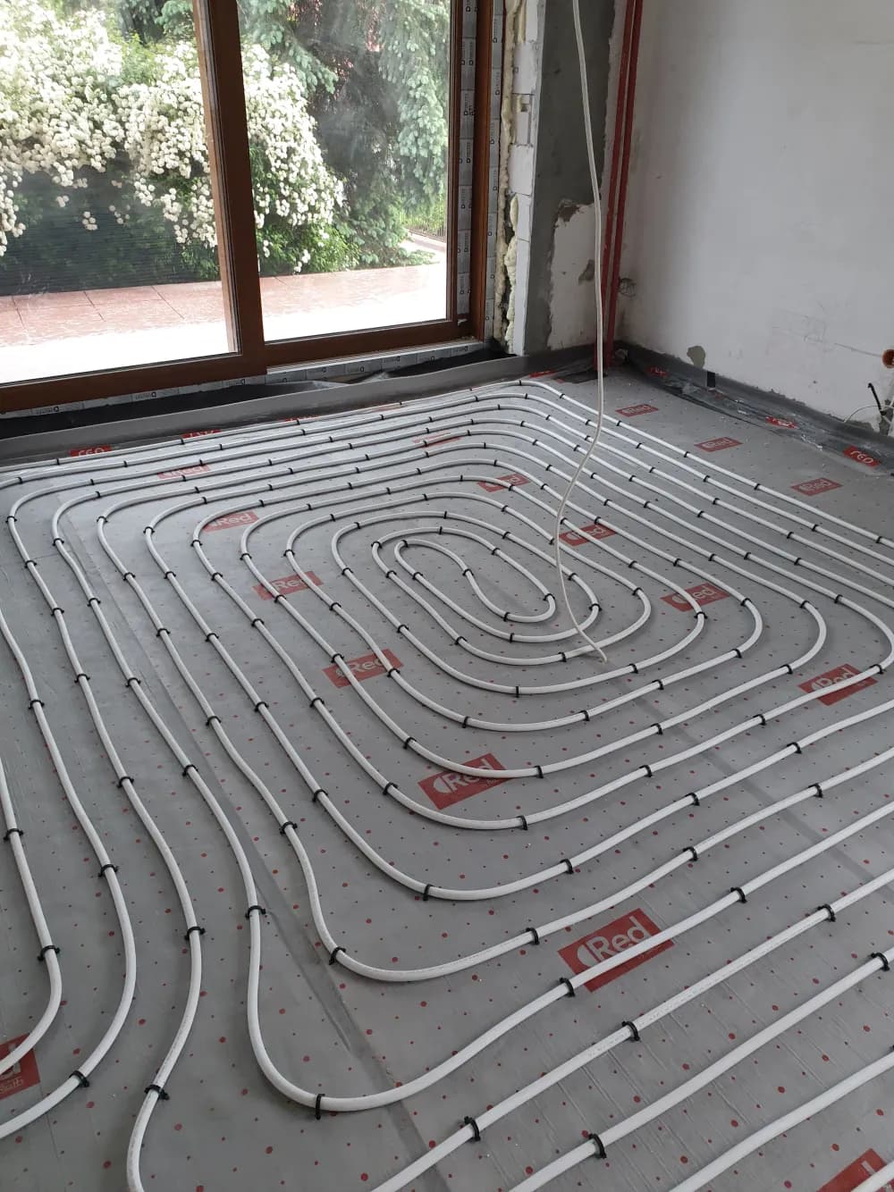Białe rury PEX ułożone spiralnie na folii z siatką w pokoju z dużym oknem tarasowym